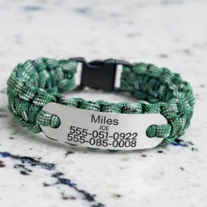 May include: A green and white paracord bracelet featuring a silver-toned metal identification plate engraved with the name Miles, the acronym ICE, and two phone numbers: 555-051-0922 and 555-085-0008, secured with a black plastic side-release buckle.