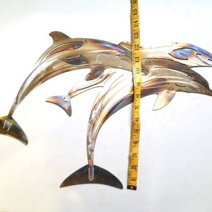 May include: Metal wall art sculpture of two dolphins jumping out of the water. The dolphins are made of brushed stainless steel with a rainbow iridescent finish. The sculpture is approximately 15 inches tall.
