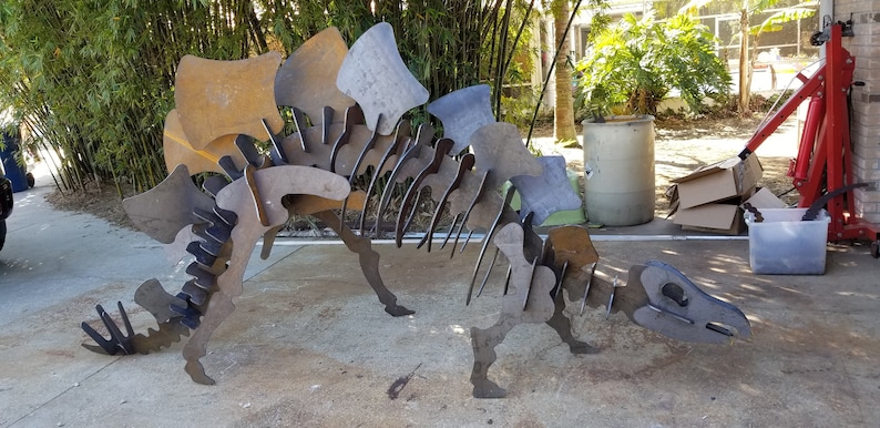 May include: A metal dinosaur sculpture made of flat pieces of metal. The sculpture is rusty in color and is standing on a concrete surface.
