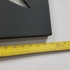 Puede incluir: Un letrero rectangular negro con una forma de media luna recortada. El letrero se mide con una regla amarilla, mostrando una longitud de 68,58 cm.