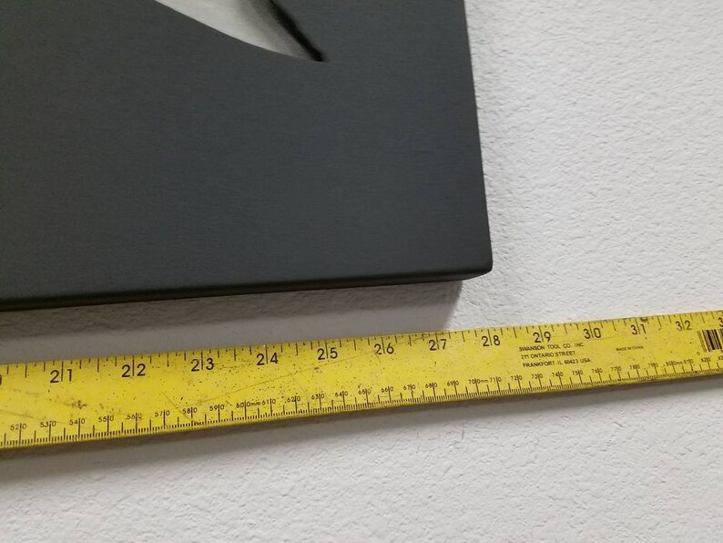 Puede incluir: Un letrero rectangular negro con una forma de media luna recortada. El letrero se mide con una regla amarilla, mostrando una longitud de 68,58 cm.