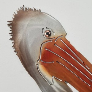 Puede incluir: Primer plano de una escultura de metal de la cabeza de un pel&iacute;cano. La escultura est&aacute; pintada en tonos de marr&oacute;n, gris y naranja. El ojo del pel&iacute;cano es un peque&ntilde;o c&iacute;rculo redondo naranja.