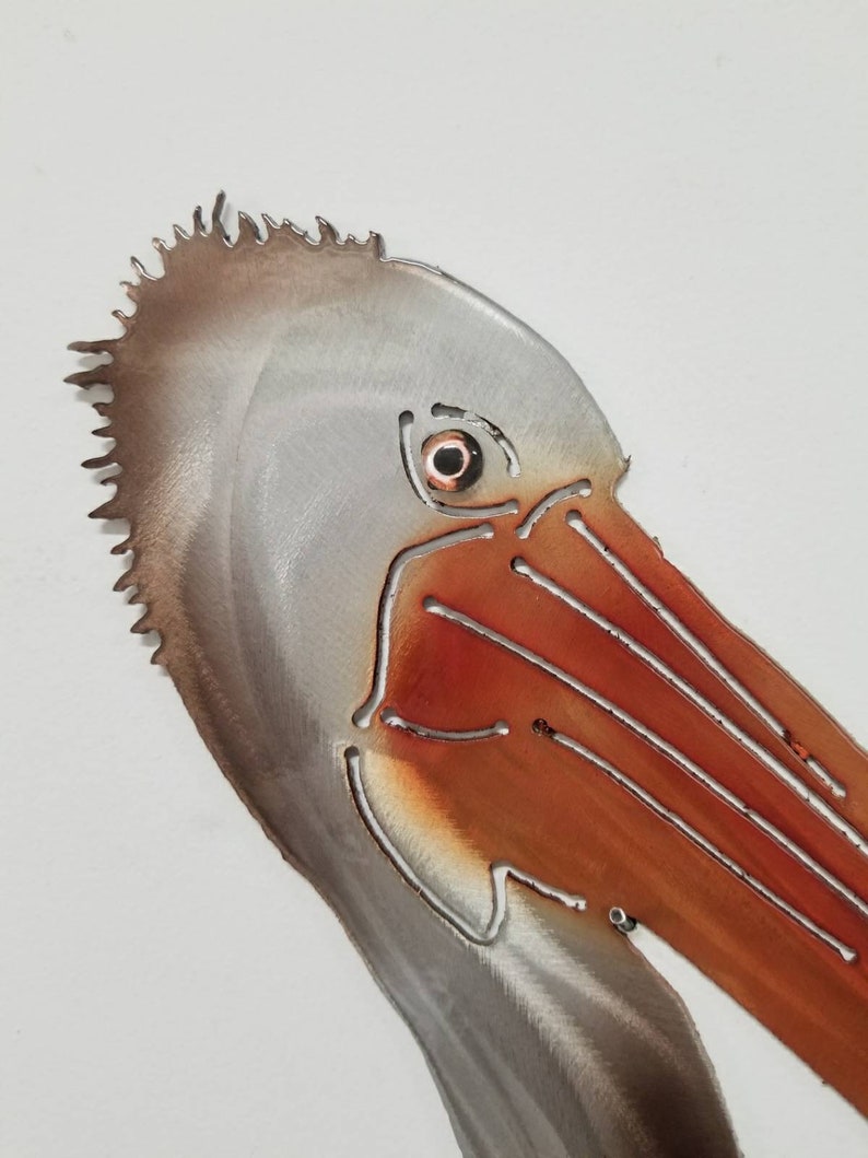 Puede incluir: Primer plano de una escultura de metal de la cabeza de un pel&iacute;cano. La escultura est&aacute; pintada en tonos de marr&oacute;n, gris y naranja. El ojo del pel&iacute;cano es un peque&ntilde;o c&iacute;rculo redondo naranja.