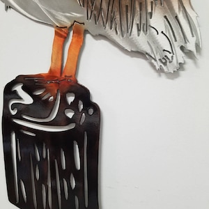 Puede incluir: Escultura de pared de metal de un pel&iacute;cano de pie sobre un toc&oacute;n de &aacute;rbol. El pel&iacute;cano est&aacute; pintado en tonos de gris, marr&oacute;n y naranja. El toc&oacute;n de &aacute;rbol est&aacute; pintado de negro.