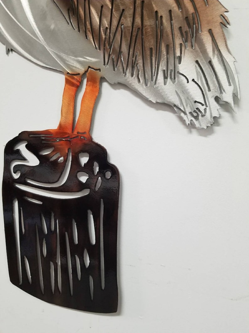 Puede incluir: Escultura de pared de metal de un pel&iacute;cano de pie sobre un toc&oacute;n de &aacute;rbol. El pel&iacute;cano est&aacute; pintado en tonos de gris, marr&oacute;n y naranja. El toc&oacute;n de &aacute;rbol est&aacute; pintado de negro.