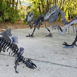 May include: Three metal dinosaur skeletons, one with wings, standing on a concrete surface. The skeletons are made of black metal and are posed in a lifelike manner.