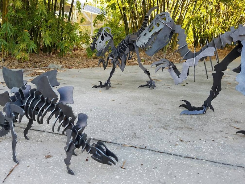 May include: Three metal dinosaur skeletons, one with wings, standing on a concrete surface. The skeletons are made of black metal and are posed in a lifelike manner.