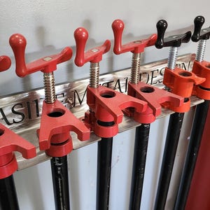 May include: A set of six red and black pipe clamps mounted on a silver metal wall rack. The rack has the text "Slash Metal Designs" printed on it.