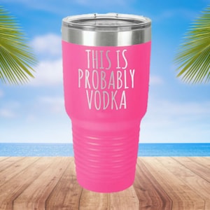 May include: A bright pink insulated tumbler with a silver lid. The text "THIS IS PROBABLY VODKA" is printed in white. The tumbler is on a wooden surface with a beach and palm trees in the background.