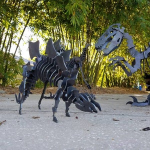 May include: Two metal dinosaur skeletons, one a Stegosaurus and the other a Tyrannosaurus Rex, are posed in a garden setting. The skeletons are made of black metal and are detailed with realistic bone structures.