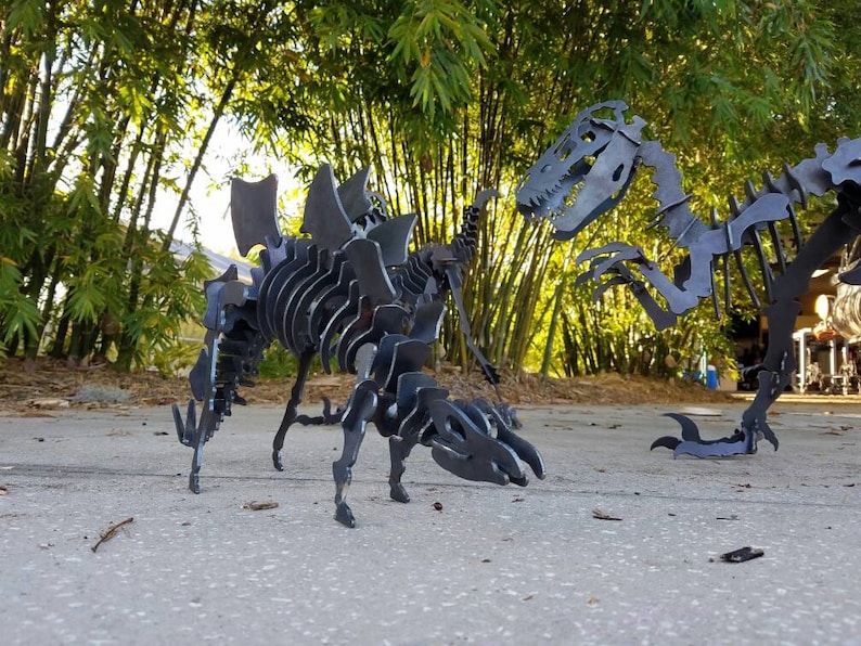 May include: Two metal dinosaur skeletons, one a Stegosaurus and the other a Tyrannosaurus Rex, are posed in a garden setting. The skeletons are made of black metal and are detailed with realistic bone structures.