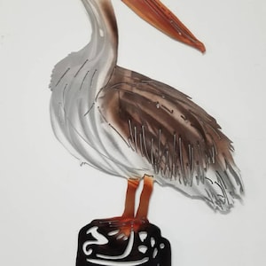 Puede incluir: Escultura de pared de metal de un pel&iacute;cano marr&oacute;n y blanco de pie sobre un toc&oacute;n de &aacute;rbol negro. El pel&iacute;cano tiene un largo pico naranja y mira hacia la derecha.
