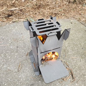 Puede incluir: Un pequeño hogar de fuego portátil de metal con una rejilla y una puerta. El hogar de fuego está sentado en una superficie de concreto y tiene un pequeño fuego ardiendo en su interior.