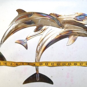 May include: Metal wall art sculpture of two dolphins jumping out of the water. The dolphins are made of brushed metal with a rainbow iridescent finish. The sculpture is approximately 20 inches wide.