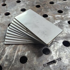 Pu&ograve; includere: Una pila di dieci lamine di metallo argentate, ciascuna con una superficie liscia e lucida, sono disposte a ventaglio su una superficie di metallo grigia con fori equidistanti.