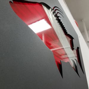 Puede incluir: Una pieza de arte de pared de metal en forma de pez con un dise&ntilde;o rojo y plateado. El pez est&aacute; cortado de una l&aacute;mina de metal negro y el dise&ntilde;o rojo y plateado est&aacute; detr&aacute;s de la l&aacute;mina de metal.