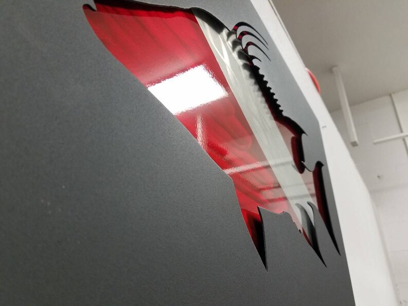 Puede incluir: Una pieza de arte de pared de metal en forma de pez con un dise&ntilde;o rojo y plateado. El pez est&aacute; cortado de una l&aacute;mina de metal negro y el dise&ntilde;o rojo y plateado est&aacute; detr&aacute;s de la l&aacute;mina de metal.