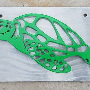May include: A green metal sea turtle cutout on a silver background. The turtle is in a swimming position with its head turned to the left. The turtle's shell is detailed with a pattern of cutouts.