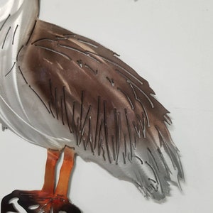 Puede incluir: Una escultura de metal de un pel&iacute;cano de pie sobre un tronco. El pel&iacute;cano est&aacute; pintado en tonos de gris, marr&oacute;n y naranja. La escultura es una pieza decorativa para un hogar o jard&iacute;n.