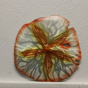 Pu&ograve; includere: Un decoro da parete a forma di dollaro di sabbia in metallo con un design dipinto di bianco, arancione e giallo. Il dollaro di sabbia ha una stella a cinque punte al centro.