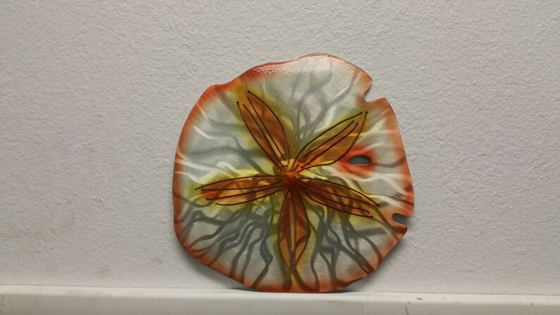 Pu&ograve; includere: Un decoro da parete a forma di dollaro di sabbia in metallo con un design dipinto di bianco, arancione e giallo. Il dollaro di sabbia ha una stella a cinque punte al centro.