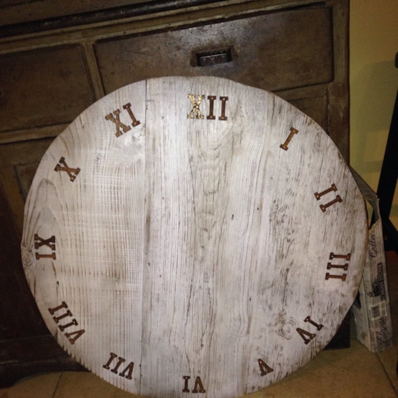 May include: A white wooden clock face with Roman numeral markings in gold. The clock face is round and has a distressed white finish.