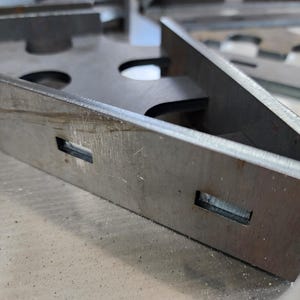 May include: Close-up of a metal bracket with two rectangular cutouts. The bracket is made of a silver-colored metal and has a rough, textured surface.