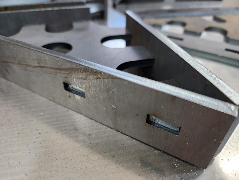 May include: Close-up of a metal bracket with two rectangular cutouts. The bracket is made of a silver-colored metal and has a rough, textured surface.