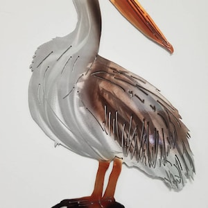 Puede incluir: Escultura de pared de metal de un pel&iacute;cano de pie sobre una roca. El pel&iacute;cano es gris con marcas marrones y tiene un pico naranja. La roca es negra.