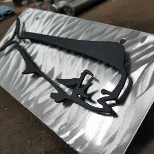 May include: A black metal silhouette of a fish, possibly a mahi-mahi, mounted on a brushed metal surface. The fish is cut out of metal and has a detailed design.