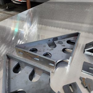 May include: Close-up of several metal brackets with cutouts. The brackets are arranged on a silver surface.