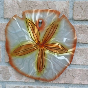 Pu&ograve; includere: Un decoro da parete a forma di dollaro di sabbia in metallo con sfondo bianco e un design a stella marina marrone.