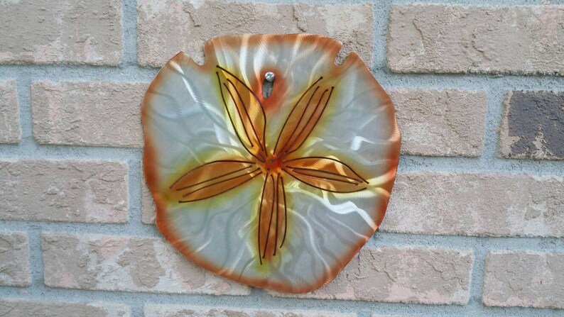 Pu&ograve; includere: Un decoro da parete a forma di dollaro di sabbia in metallo con sfondo bianco e un design a stella marina marrone.