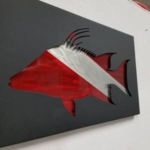 Puede incluir: Una escultura de pared de metal de un pez con un dise&ntilde;o de bandera de buceo roja y blanca. El pez est&aacute; cortado del metal y la bandera est&aacute; detr&aacute;s.