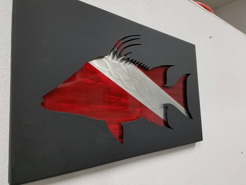 Puede incluir: Una escultura de pared de metal de un pez con un dise&ntilde;o de bandera de buceo roja y blanca. El pez est&aacute; cortado del metal y la bandera est&aacute; detr&aacute;s.
