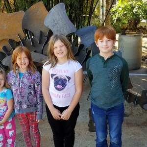 May include: Four children stand in front of a large metal sculpture. The sculpture is made of many pieces of metal that are welded together. The children are all smiling and looking at the camera. The girl in the middle is wearing a white shirt with a pug on it and the text "This is my birthday face".
