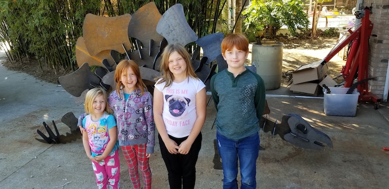 May include: Four children stand in front of a large metal sculpture. The sculpture is made of many pieces of metal that are welded together. The children are all smiling and looking at the camera. The girl in the middle is wearing a white shirt with a pug on it and the text "This is my birthday face".