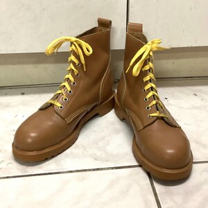 Puede incluir: Un par de botas de cuero marrón con cordones amarillos. Las botas tienen un diseño clásico con punta redonda y suela resistente. Los cordones están atados con un lazo en la parte superior. Las botas son de color marrón medio.