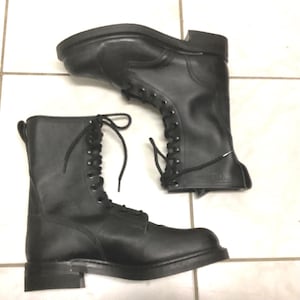 May include: A pair of black leather combat boots with lace-up fastenings. The boots have a robust design with a thick sole and a tall shaft. The boots are displayed on a tiled surface.