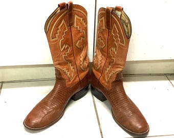 May include: A pair of brown cowboy boots with intricate stitching and a textured design on the foot. The boots have a classic western style with a pointed toe and a stacked heel. The shaft of the boots is a lighter shade of brown with decorative stitching.