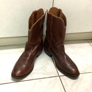 Puede incluir: Un par de botas de vaquero de cuero marrón. Las botas tienen un diseño clásico con puntas puntiagudas, tacones angulados y presillas. El cuero tiene un tono rico y cálido, y las botas parecen gastadas.