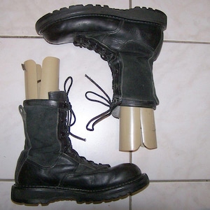 Puede incluir: Un par de botas tácticas negras de cuero y tela con cordones negros y suelas de goma gruesas. Cada bota contiene un inserto moldeador de plástico beige para mantener la forma de la caña y el área del tobillo.