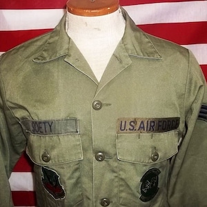 Peut inclure: Chemise d'uniforme vert olive de l'US Air Force. La chemise a deux poches de poitrine, des boutons et des écussons. Le nom "SOETY" est brodé sur une étiquette nominative au-dessus de la poche gauche. Les mots "U.S. AIR FORCE" sont sur un écusson au-dessus de la poche droite.