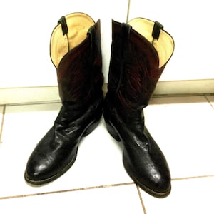 May include: A pair of black leather cowboy boots featuring a textured finish on the foot and a dark red shaft with black stitching details. The boots have a pointed toe, a low heel, and a light-coloured interior lining with black pull tabs.