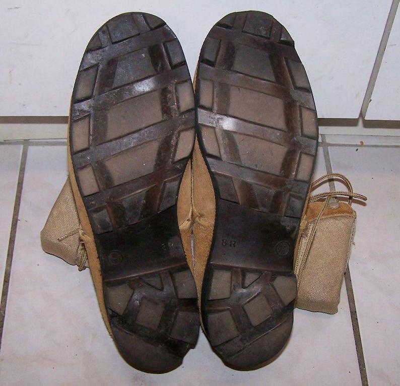 K&ouml;nnte beinhalten: Nahaufnahme der Sohlen eines Paares brauner Lederstiefel mit einem braunen Gummibrockenmuster. Die Stiefel haben ein braunes Leder-Obermaterial und einen beigefarbenen Canvas-Kn&ouml;chelriemen. Die Gr&ouml;&szlig;e "8R" ist auf der Sohle des rechten Stiefels aufgedruckt.