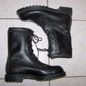 Puede incluir: Un par de botas de combate de cuero negro con diseño de caña alta, suelas de goma gruesas con tacos y cordones negros. Las botas tienen un acabado liso con detalles de costuras visibles y ojales metálicos para el sistema de cordones.