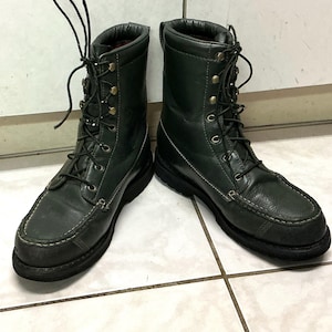 Puede incluir: Un par de botas de cuero verde oscuro con puntera estilo mocasín, suelas gruesas de goma negra y un diseño de caña alta con cordones, ojales y ganchos metálicos. Las botas tienen costuras visibles de color claro a lo largo de los bordes.