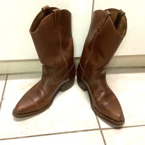 Puede incluir: Un par de botas de vaquero de cuero marrón. Las botas tienen una punta afilada, un tacón de altura media y presillas en la parte superior. El cuero tiene un aspecto ligeramente desgastado. Las botas están sobre un fondo de baldosas blancas.