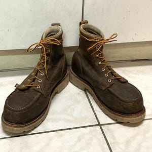 May include: A pair of dark brown leather work boots featuring a moccasin-style toe, thick light-coloured rubber lug soles, and yellow and orange braided laces threaded through silver-toned metal eyelets and hooks at the ankle collar.