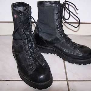 May include: A pair of black leather combat boots with a lace-up design. The boots have a high-top silhouette, a textured sole, and a fabric panel on the upper shaft. An American flag patch is visible on the side of one boot.